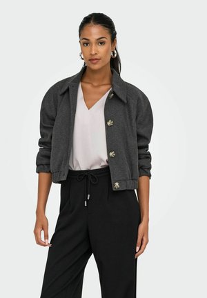 Naine seisab enesekindlalt, seljas hall lühikeste varukatega jakk, kerge blouse ja mustad püksid, kõrvarõngad ja sirged, tagasi kammitud juuksed.