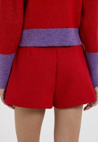 Maglione lavorato a maglia rosso con bordi viola alle maniche, abbinato a pantaloni corti rossi. Il design presenta una vestibilità comoda e dettagli di cucitura visibili.