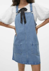 Kvinna i en blå denimoverallklänning med framfickor och justerbara axelremmar över en vit texturerad blus med puffärmar och en svart rosett.