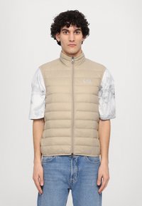 Beige oppustet vest med høy krage og horisontale sømmer. Pyntet med logo på brystet. Brukt over en lys, marmorert T-skjorte med korte ermer.
