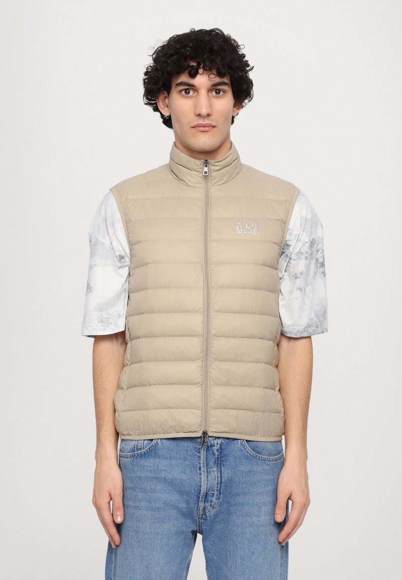 Beige oppustet vest med høy krage og horisontale sømmer. Pyntet med logo på brystet. Brukt over en lys, marmorert T-skjorte med korte ermer.