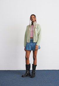 Veste en polaire vert clair, crop top rose, mini jupe en denim et bottes noires jusqu'au genou ; présente des textures et des éléments de design superposés.