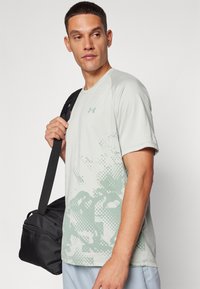 T-shirt sportivo grigio chiaro con un motivo astratto verde. Maniche corte, colletto rotondo e logo sul petto. Il modello tiene una borsa nera.