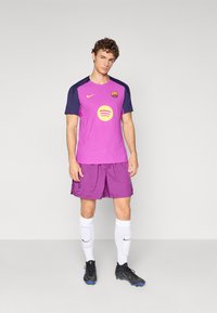 Lilla fotbollsdräkt med en rund gul logga, marinblå ärmar, matchande lila shorts, vita strumpor och svarta fotbollsskor.