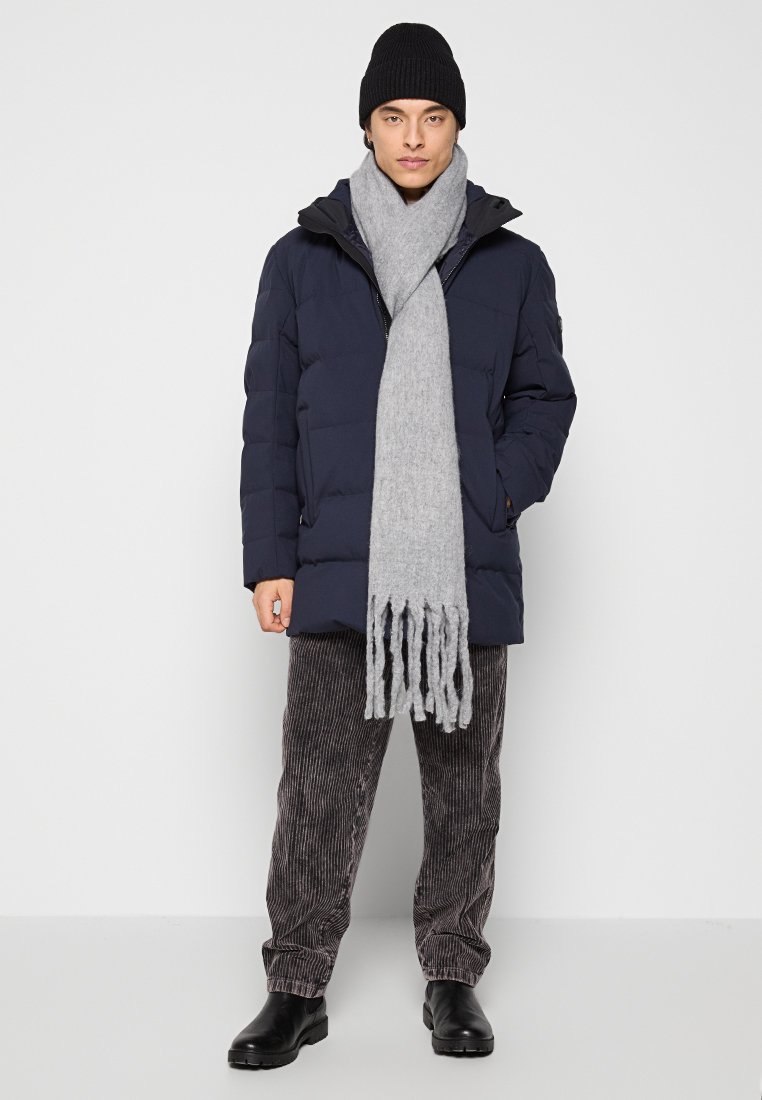 Marineblaue Daunenjacke, grauer Strickschal mit Fransen, Cordhosen, schwarze Stiefel und Mütze, alles in einer neutralen Umgebung.