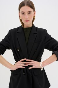 Juodas dvigubas švarkas su išsiskiriančiais mygtukais ir kišenėmis, uždėtas ant tamsiai žalios megztinio. Modelis nešioja perlų apyrankę.