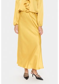Donna che indossa una lunga gonna di raso gialla e una blusa coordinata con scollo a rouches, abbinata a décolleté con punta nera e cinturino.