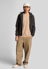 Giacca nera leggera da uomo con cappuccio, t-shirt beige, pantaloni khaki e sneakers crema. Tessuto liscio, vestibilità relax e colori neutri.