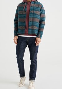 Mönstrad flanellskjorta i flera färger med geometriskt mönster, cyan- och bruna nyanser, två bröstfickor, lager på en vinröd hoodie, kombinerad med mörka jeans.