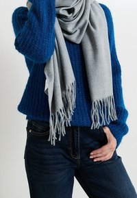 Écharpe grise à franges drapée sur un pull tricoté bleu texturé, associée à un jean en denim foncé.