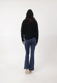 Suéter negro de punto, jeans acampanados azules y tacones color beige. El cabello es largo y ondulado, visto desde atrás contra un fondo blanco sencillo.