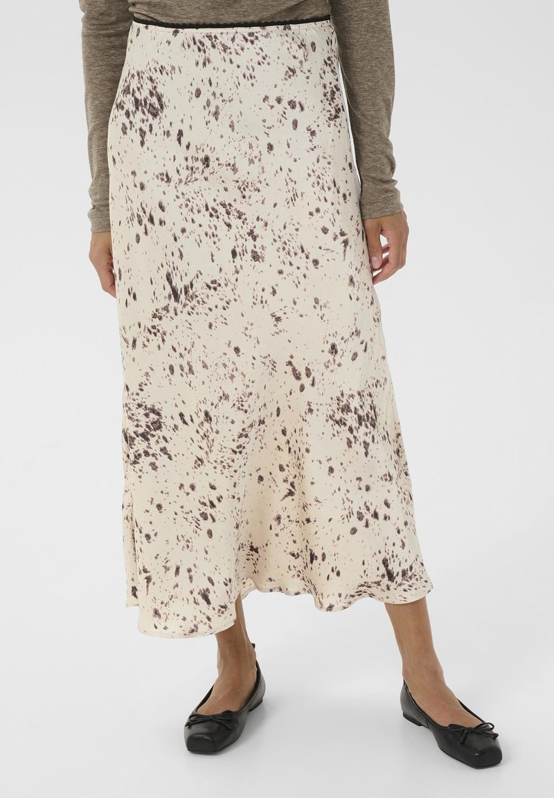 Femme portant une jupe midi crème avec un motif tacheté marron foncé, associée à un haut marron à manches longues et des ballerines noires.