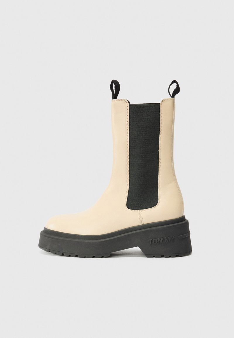 Bottines beiges avec un panneau latéral élastique noir et une semelle en caoutchouc, présentant un logo de marque bien en vue sur le talon. Design simple et moderne.