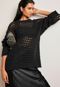 Pulover negru tricotat cu un design lejer, asemănător unui plasă, mâneci largi și manșete fibrilate, purtat peste o fustă de satin închis. Include o geantă mică.