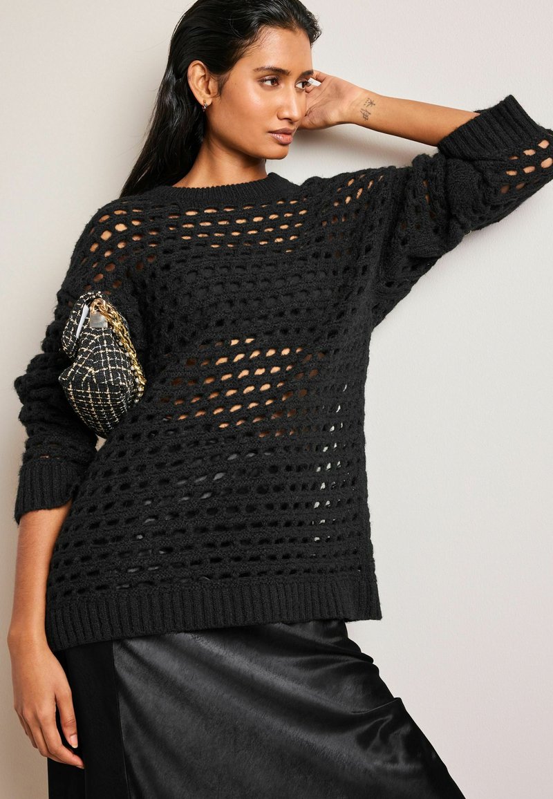Pulover negru tricotat cu un design lejer, asemănător unui plasă, mâneci largi și manșete fibrilate, purtat peste o fustă de satin închis. Include o geantă mică.