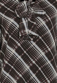 Brun och vit mönstrad blus med en rosett vid halsen; har diagonal rutigt mönster och mjuk struktur.