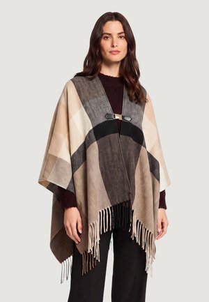 Donna che indossa un poncho a frange beige e marrone a quadri, fissato con una fibbia nera e dorata sopra un maglione scuro e pantaloni neri.