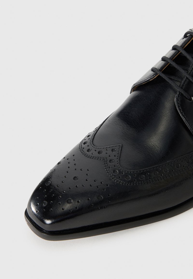 Chaussure habillée en cuir noir avec des détails brogues et des lacets sur un fond blanc, mettant en avant la partie pointue de la chaussure.