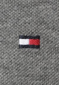 Harmaa neulekangas, jossa on pieni brodeerattu logo punaisilla, valkoisilla ja sinisillä raidoilla, sijaitsee sivussa.