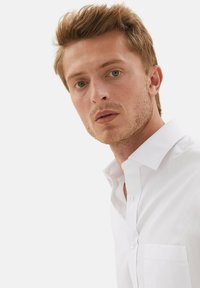 Camisa blanca de botones confeccionada con tela suave, con cuello puntiagudo y un único bolsillo en el pecho. Ajuste limpio y entallado, sin patrones visibles.