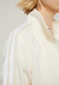Lys beige zip-jakke med hvite striper langs skuldrene, glatt tekstur og høy krage. Minimalistisk design uten synlige logoer.