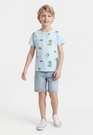 Αγόρι που στέκεται και χαμογελάει, φορώντας ανοιχτό μπλε μπλούζα με σχέδιο βατράχου, τζιν σορτς και λευκά αθλητικά παπούτσια, σε λευκό φόντο.