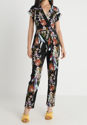 Combinaison fleurie en tissu noir à manches courtes, design cache-cœur, taille ceinturée, avec des motifs vifs de roses et de feuilles. Portée avec des sandales jaunes.