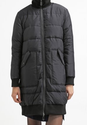 Manteau d'hiver matelassé noir longueur genou avec fermeture éclair frontale, deux poches à rabat, poignets et ourlet côtelés, porté par une personne.