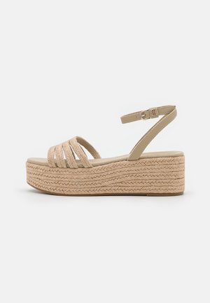 Beige platform sandal med flettet sål, flere frontremme og justerbar ankelrem med metalspænde på en hvid baggrund.