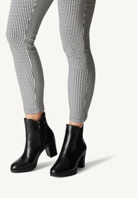 Bottines en cuir noir avec un talon bloc haut et une fermeture éclair sur le côté, associées à un pantalon ajusté à carreaux noir et blanc.