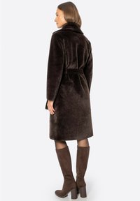 Abrigo de piel sintética marrón con cintura ajustada, que presenta un cuello alto y un diseño de longitud media. Combinado con botas de tacón marrones hasta la rodilla.