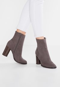 Botas de tobillo de ante gris con un tacón cuadrado, cremallera lateral, tacón de textura similar a la madera y un diseño elegante y aerodinámico.