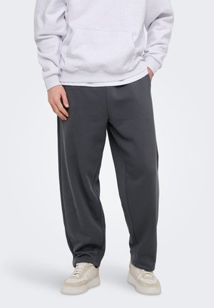 Oseba, ki nosi temno sive široke hlače, bele športne copate in svetlo sivo kapuco, stoji z rokami v žepih pred enobarvnim ozadjem.