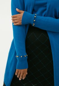 Maglione blu con polsini a coste e dettagli in perle d'oro, abbinato a una gonna nera con un motivo a diamante e dettagli luccicanti.