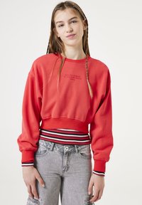 Sudadera roja recortada con mangas largas, puños acanalados y una cinturilla a rayas. Presenta texto bordado en el centro. Llevada con vaqueros grises.