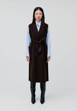 Donkerbruine wollen vestjurk met een ceintuur in de taille, twee voorzakken en een gladde textuur, gecombineerd met een lichtblauw overhemd met knopen en zwarte kniehoge laarzen.