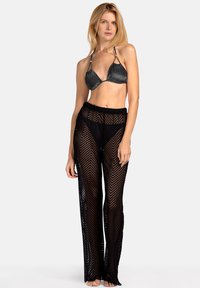 Czarne szydełkowe spodnie plażowe z wzorem w zygzak, w połączeniu z błyszczącym metalicznym bikini w kształcie trójkąta. Oba elementy są dopasowane.