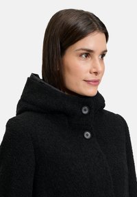 Zwarte gewatteerde jas met een textuur en een fuzzy oppervlak. Bevat twee knopen aan de voorkant en een hoge kraag voor extra warmte.