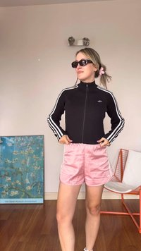 Black ribbed zip-up jacket with white stripes, paired with light pink satin shorts. Black sunglasses and a pink hair accessory are also visible.