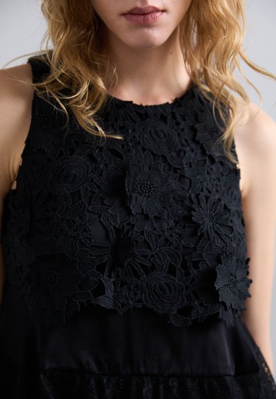 Mustsõrmevaba must särk, millel on keeruline lilleline pitsi kate, tekstuuriga riie, mis kontrastib siledaga satiiniga, ja kihiline disainiefekt.