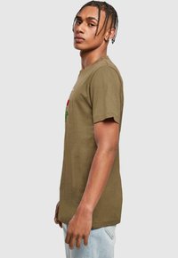 Olive green cotton t-shirt with a crew neck, short sleeves, and floral embroidery in red and green. Worn with light blue jeans.
