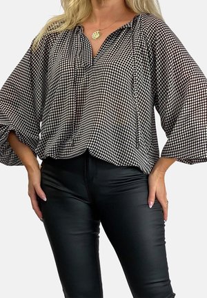 Kobieta w luźnej czarno-białej koszuli w kratę o szerokich rękawach i dopasowanych czarnych skórzanych spodniach, z rękami na biodrach.