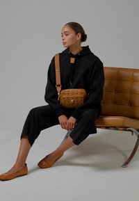 Femme assise sur une chaise en cuir beige, portant un sweat à capuche noir, un pantalon noir, des ballerines beiges et un sac bandoulière beige.