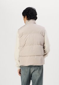 Lichtbeige puffer vest met een hoge kraag en horizontale stiksels, gedragen over een crèmekleurige trui en gecombineerd met blauwe jeans.