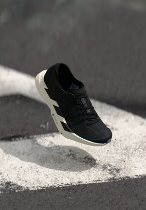 Schwarzer Sportschuh mit weißer Zickzacksohle, die über Asphalt mit weißen Fahrbahnmarkierungen und verstreutem Schmutz schwebt.