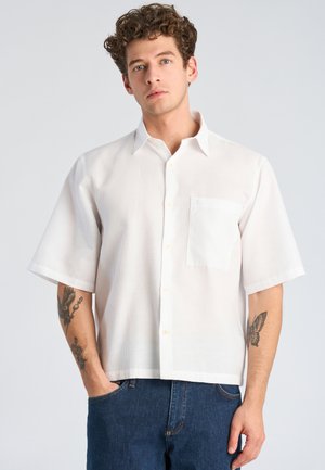 Giovane uomo con capelli ricci che indossa una camicia bianca a maniche corte con bottoni e jeans blu, con tatuaggi visibili su entrambi gli avambracci.