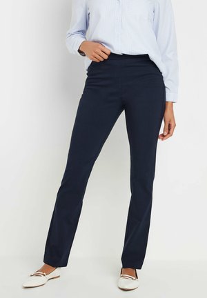 Femme portant un pantalon droit ajusté bleu marine foncé, une chemise à rayures bleu clair et des chaussures plates blanches, debout devant un fond uni.