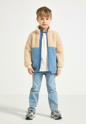 Fleecejakke i beige og blå, lynlåslukning foran, høj krave, blød tekstur, kombineret med lyse denimjeans og flerfarvede sneakers.