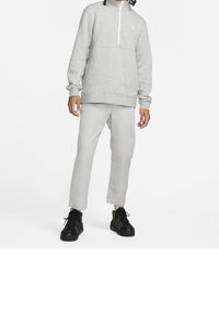 Sudadera gris con media cremallera, puños y cinturilla acanalados, combinada con pantalones gris claro y zapatillas deportivas negras con detalles verdes.