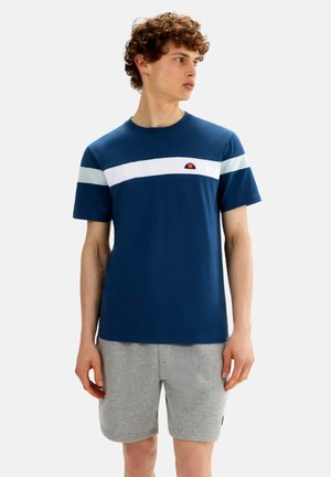 Giovane uomo con capelli ricci che indossa una t-shirt blu con strisce bianche e pantaloncini grigio chiaro, che guarda di lato su uno sfondo uniforme.